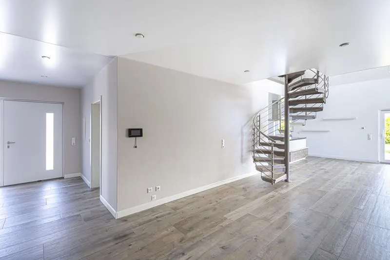 Modern open-plan room with light wood floors, white walls, a white entry door, and a metal spiral staircase.