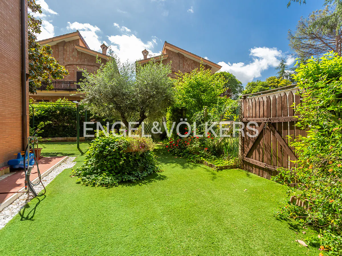 A lush green backyard with trees, flowers, and a wooden gate under a blue sky with clouds. The Engel & Völkers logo is visible.