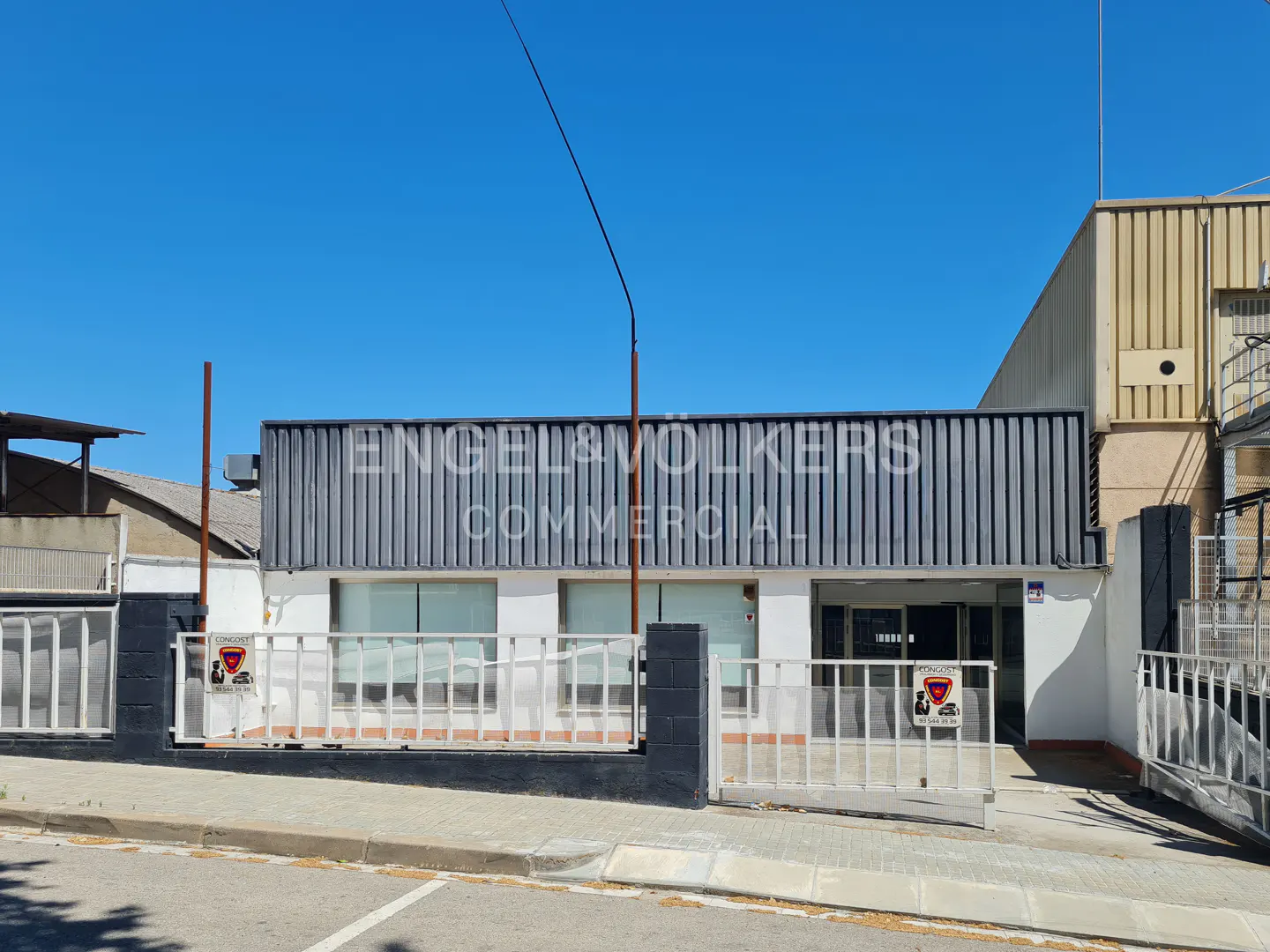 Exterior view of a commercial building with a grey corrugated metal facade, white walls, and a gated entrance under blue sky.