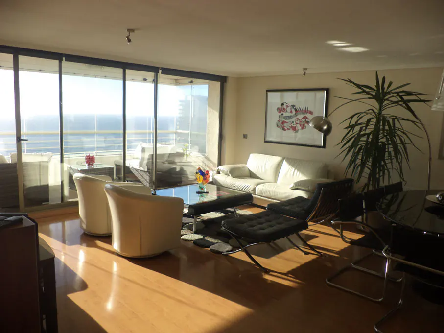 Bright living room with ocean view. White sofa and chairs, black lounge chair, and a glass coffee table. Large windows and a balcony.