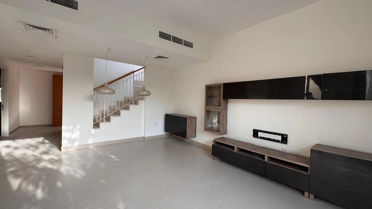 Bright, empty living room with white walls, tile floor, and dark wood entertainment center. Stairs with white railings lead up. Two pendant lights hang from the ceiling.