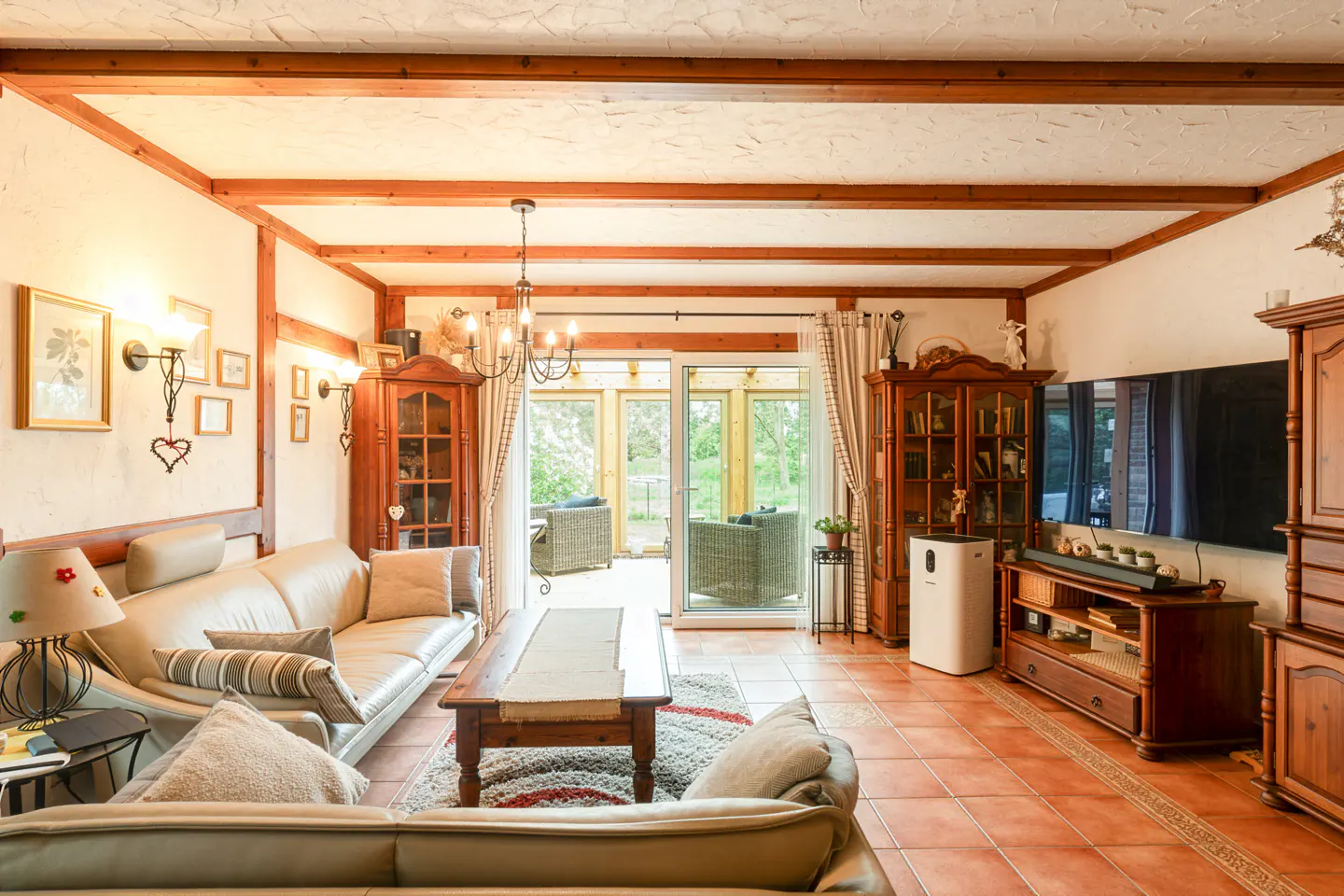 Living room with tan leather sofas, wood beams, cabinets, and terracotta tile floor. Sliding glass doors lead to an outdoor patio.