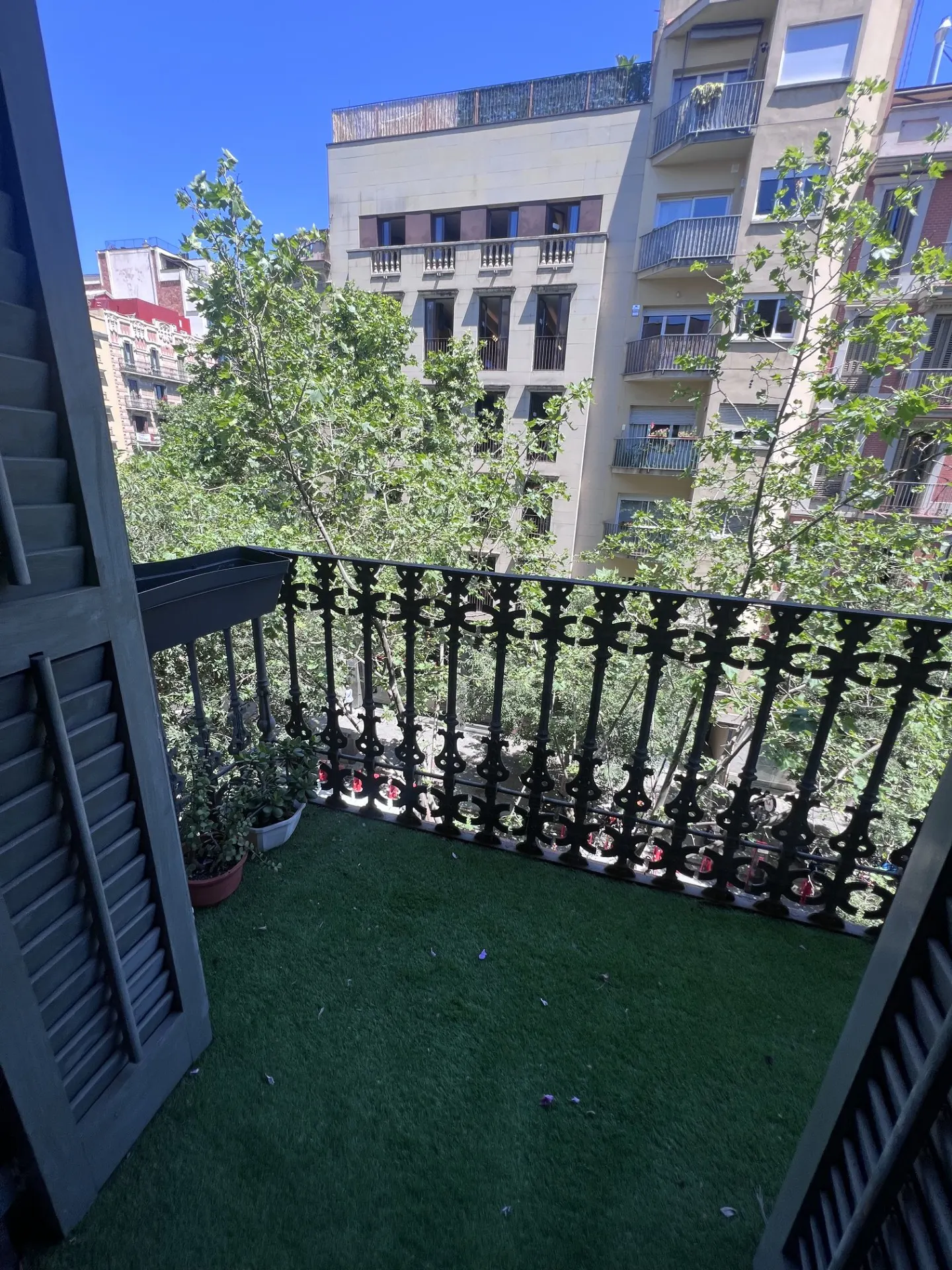 A balcony with artificial grass, a black iron railing, and green shutters. Buildings and trees are visible in the background.