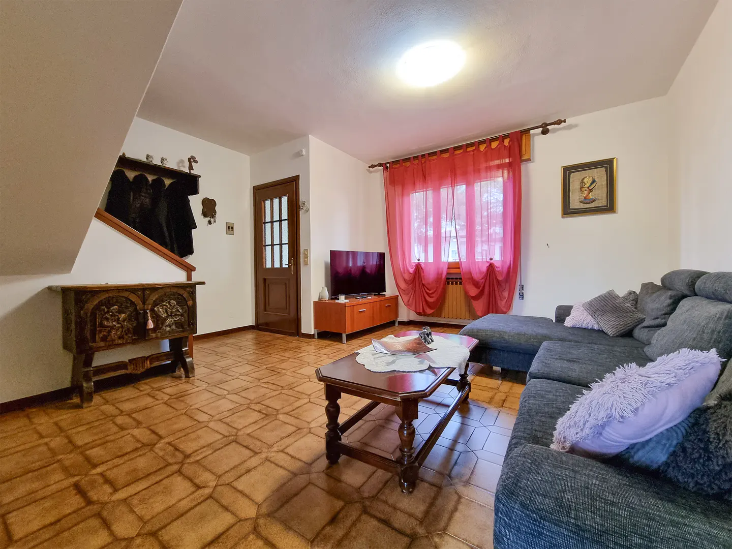 Living room with brown tile floor, gray sectional sofa, wood coffee table, TV, and red curtains.