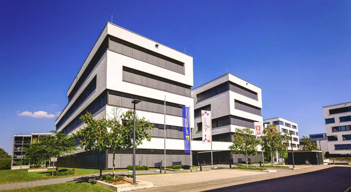 Modern white office buildings under a clear blue sky. Flags and trees line the entrance.