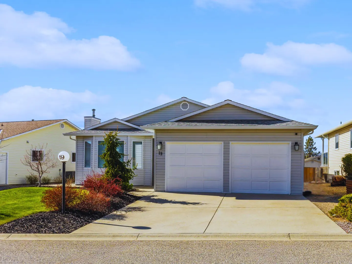 A light gray house with a two-car garage, a concrete driveway, and a lawn with red bushes.