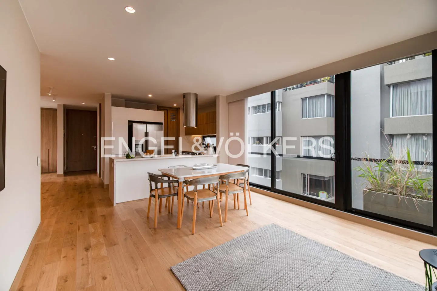 Bright, modern apartment with wood floors, a dining table with six chairs, and a large window overlooking a courtyard. Engel & Völkers logo visible.