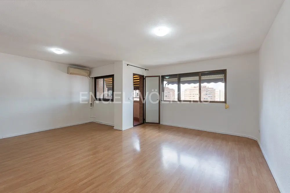 Bright, empty room with wood floors, white walls, and two windows showing city views. A doorway leads to another room.