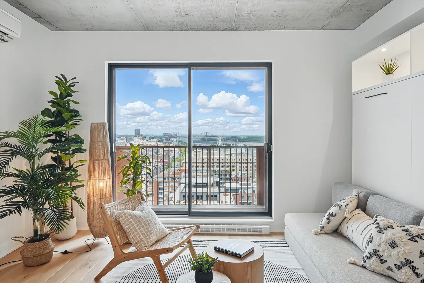 Modern living room with a grey sofa, wooden chair, floor lamp, and large window showing a city skyline view.