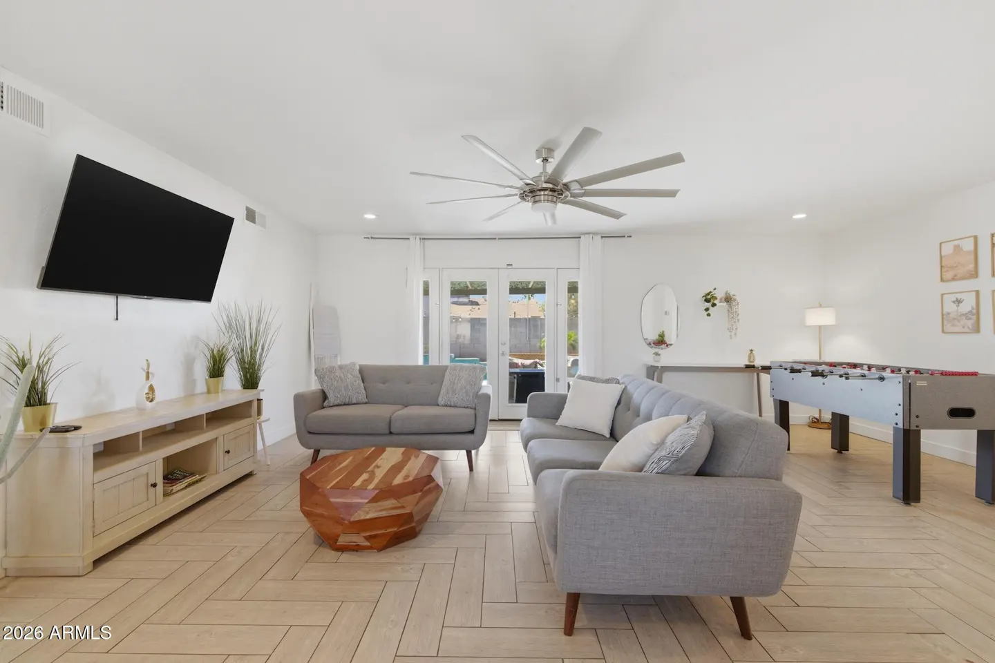 Bright living room with gray sofas, wood floors, and a foosball table. A TV hangs above a light wood console. Patio doors lead to a pool.