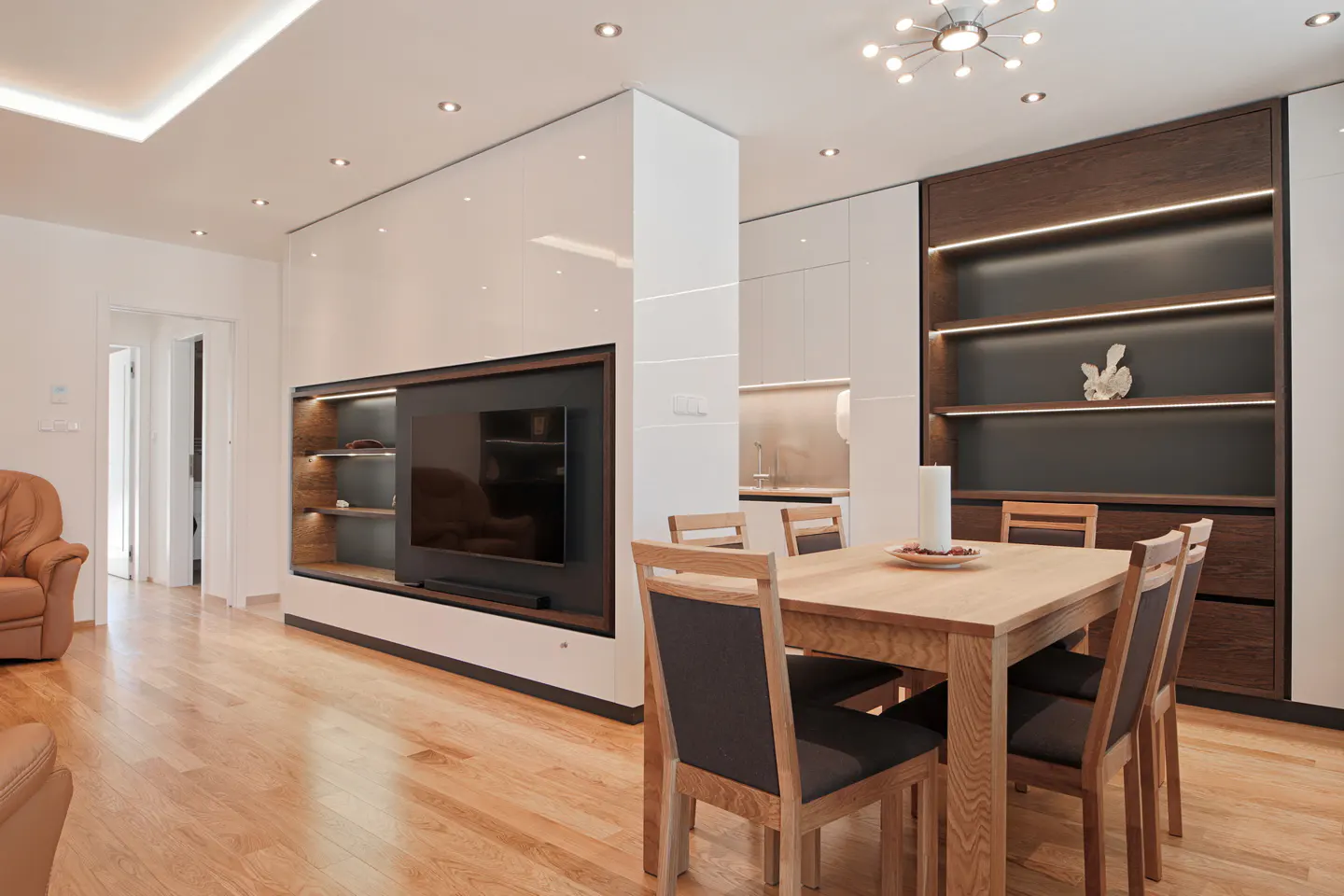 A modern dining area with a light wood table and six chairs. A TV is mounted in a white wall unit with dark wood shelving. Hardwood floors and recessed lighting.