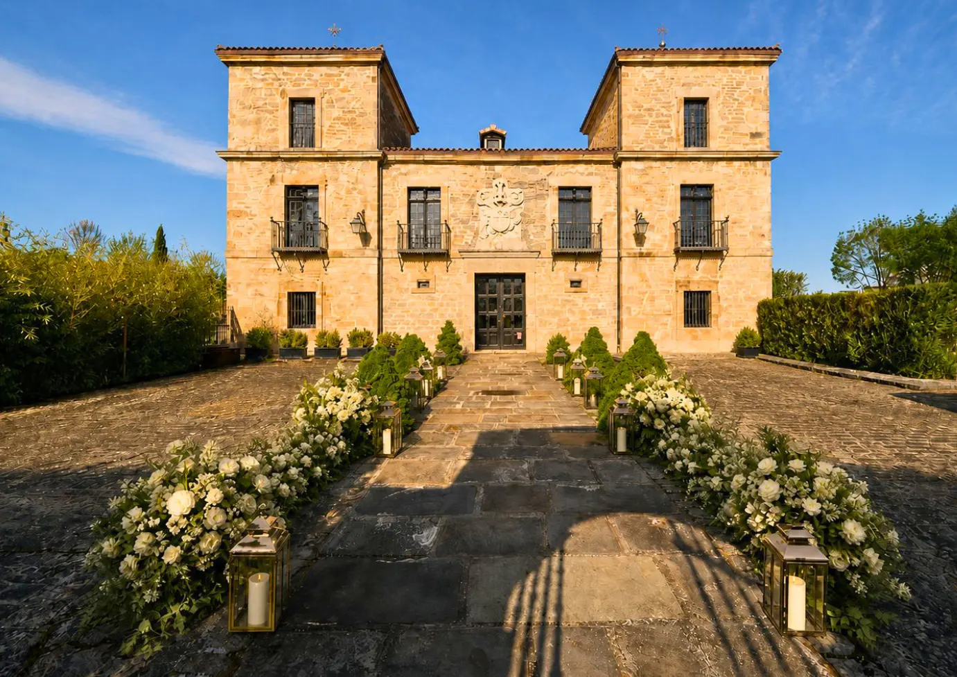 Stone mansion with two towers, under a blue sky. A stone path leads to the front door, lined with flowers and lanterns.