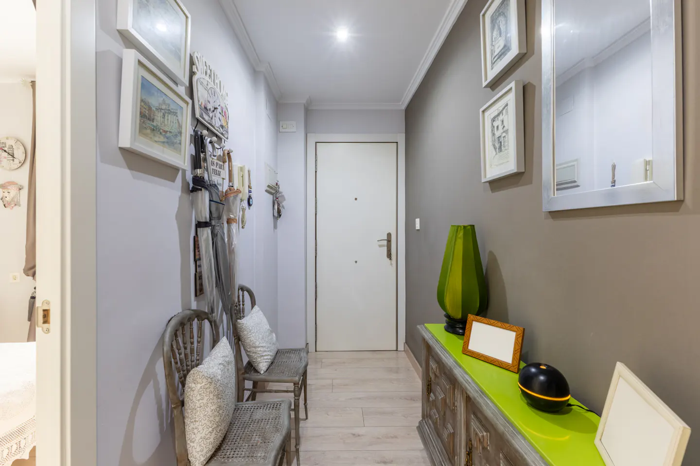Hallway with gray walls, light wood floors, and white door. Two chairs with pillows sit on the left, and a green console table is on the right.