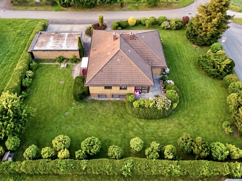 Aerial view of a tan brick house with a brown tile roof, green lawn, and trimmed hedges. A detached garage is on the left.