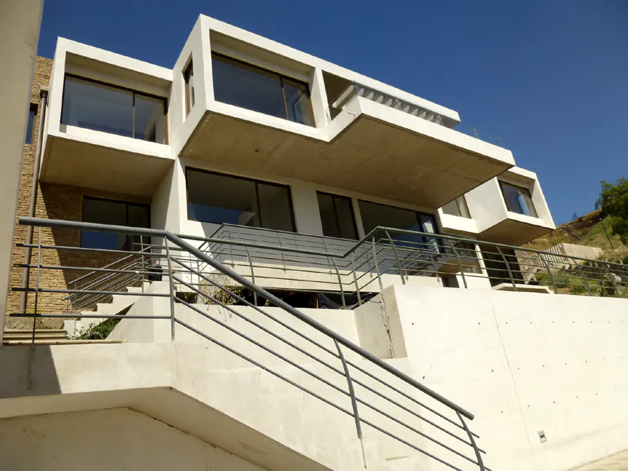 Modern white house with large windows and a gray metal railing on a hillside under a clear blue sky.