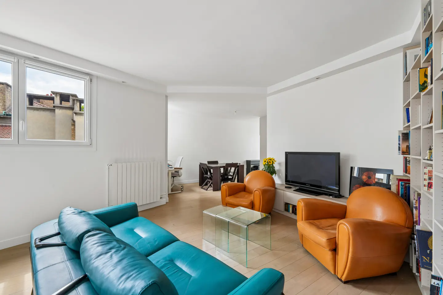 Bright living room with a blue sofa, two orange armchairs, a glass table, and a bookshelf. A window shows rooftops outside.