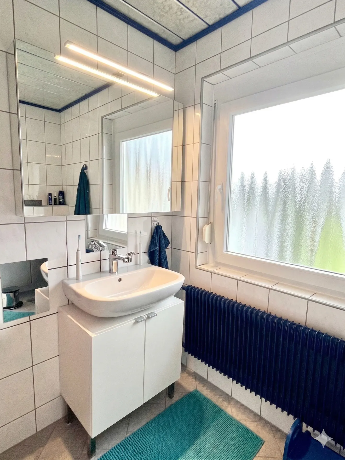 Bright bathroom with white tiled walls, a white vanity with a vessel sink, and a blue radiator under a window.