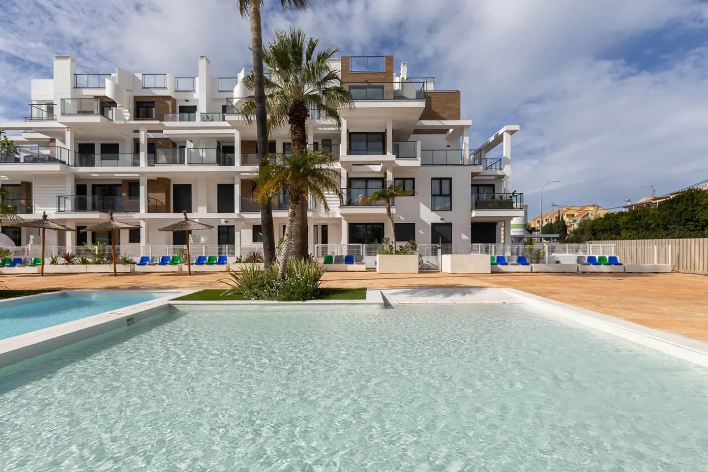 Modern white apartment building with balconies overlooks a pool. Palm trees and lounge chairs add to the resort-style atmosphere.