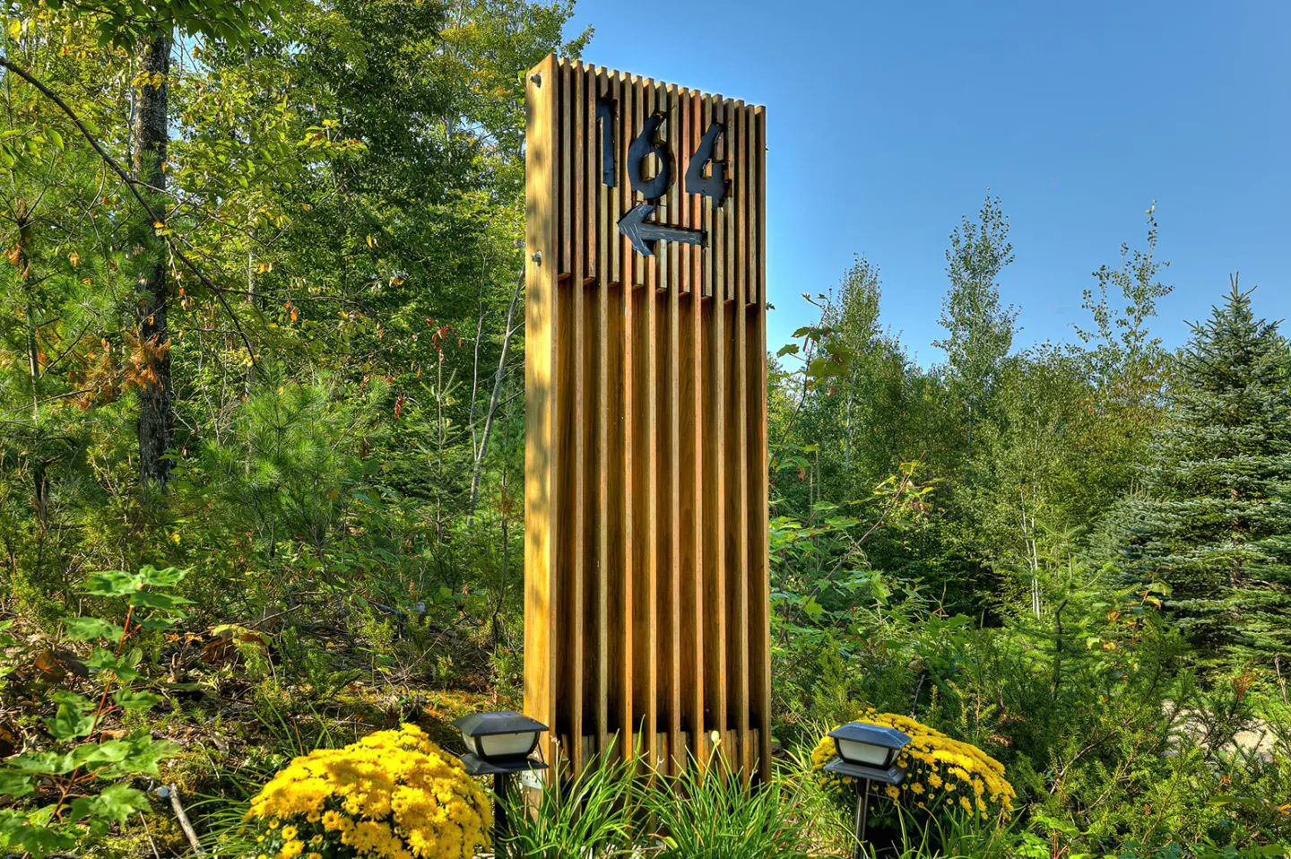 Wooden sign with address "164" and arrow, surrounded by green foliage and yellow flowers. Blue sky in the background.