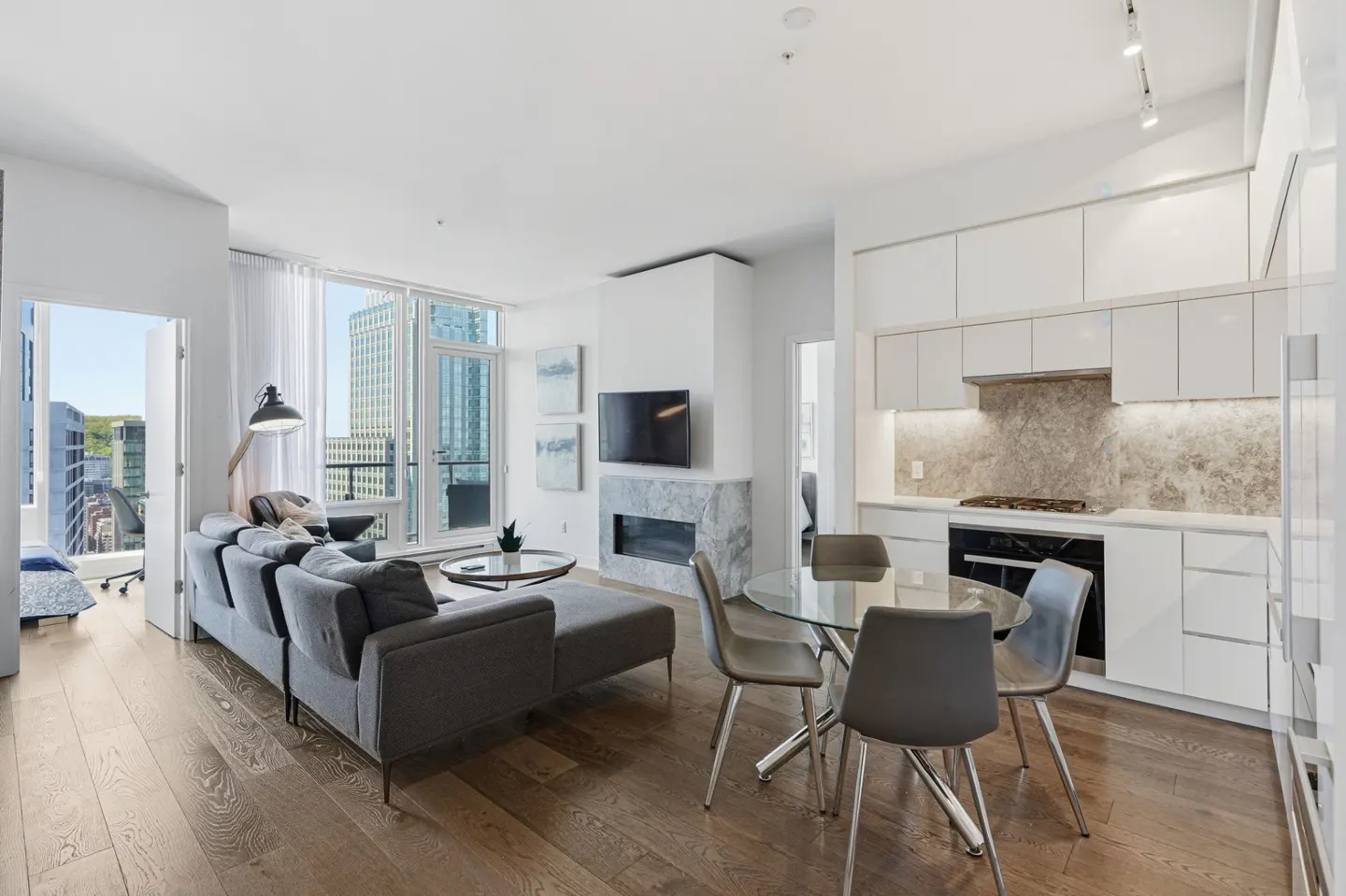Bright, modern condo interior with a gray sofa, glass table, and white kitchen. A city view is visible through the open balcony door.