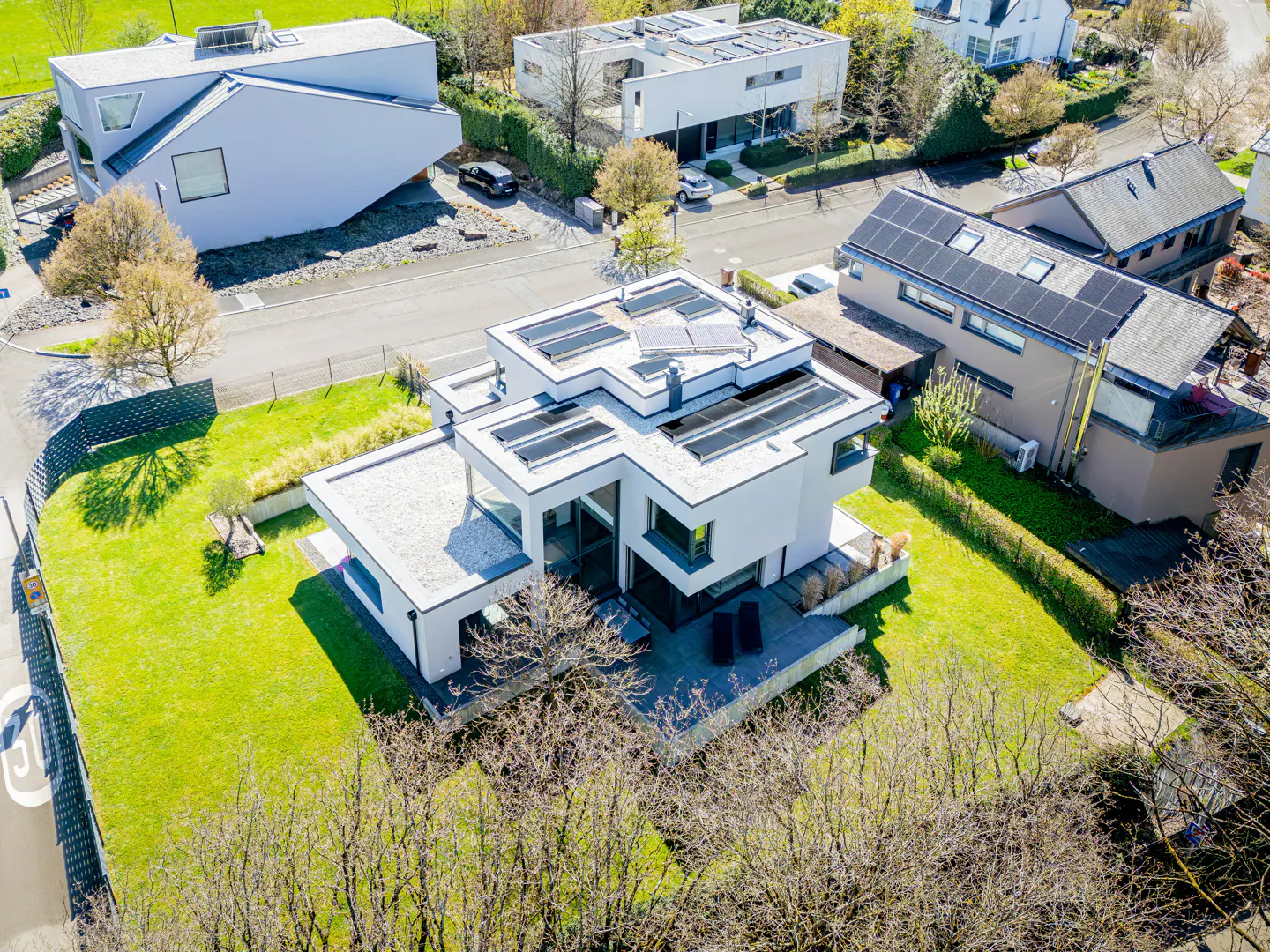 Aerial view of a modern white house with solar panels, surrounded by green lawns and trees.