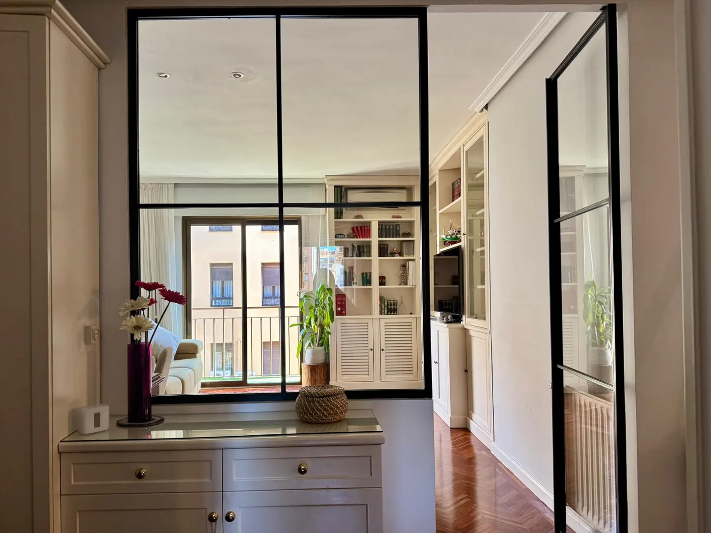 Interior view through a black-framed glass partition. A white cabinet with flowers sits in front, with a living room and balcony visible beyond.