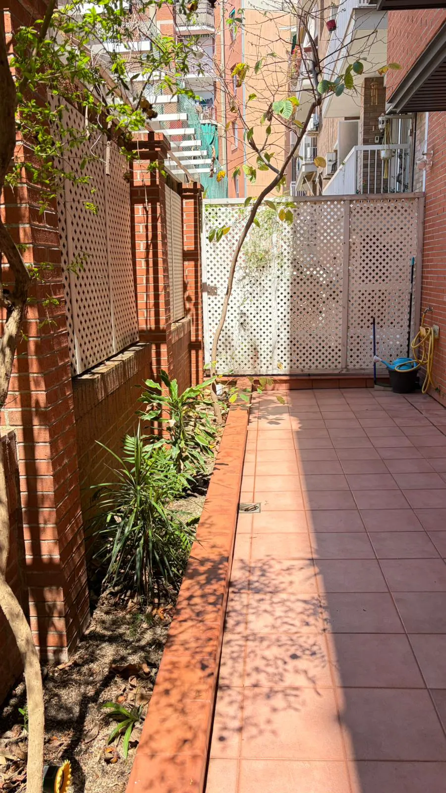 A sunny patio with red brick walls, white lattice fencing, and terracotta tiles. A small garden bed with green plants runs along the wall.
