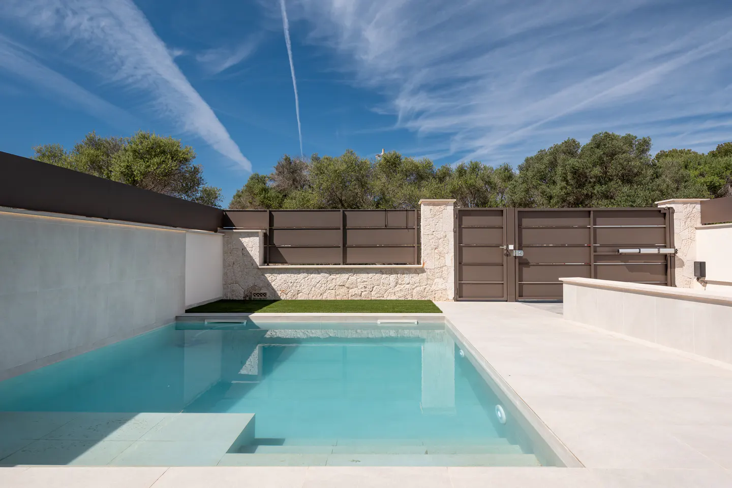 A modern rectangular pool with turquoise water, surrounded by white stone, a brown fence, and a gate under a blue sky.