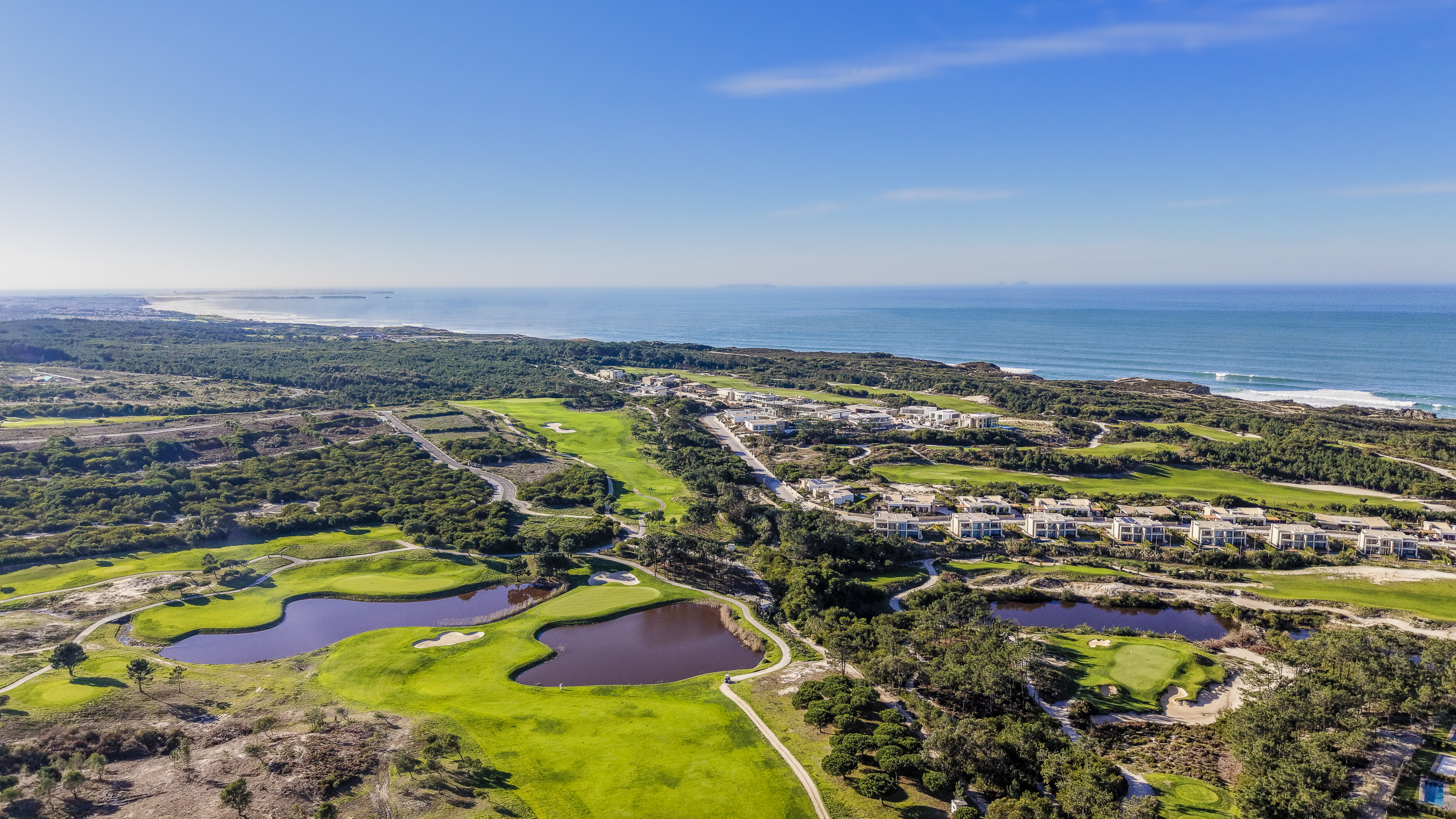 Aerial view of a golf course with ponds, green grass, and modern houses near the ocean under a blue sky.