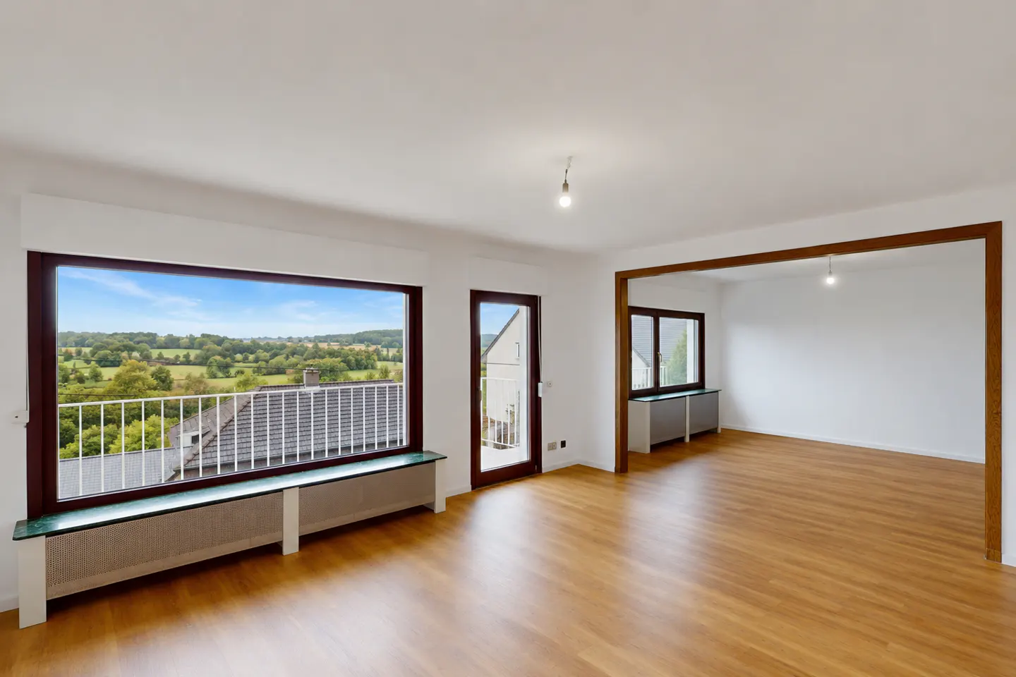 Bright, empty room with wood floors, white walls, and large windows. A balcony overlooks a green landscape. A doorway leads to another room.