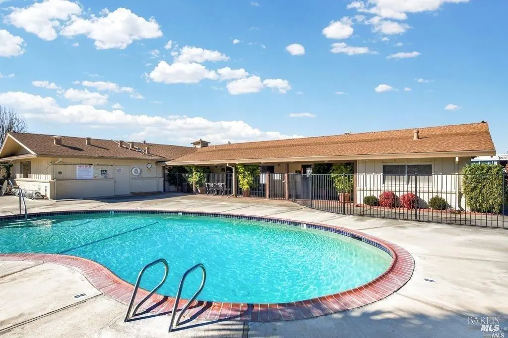 Outdoor pool with blue water and metal ladder, surrounded by a concrete deck and a building with a brown roof under a blue sky.