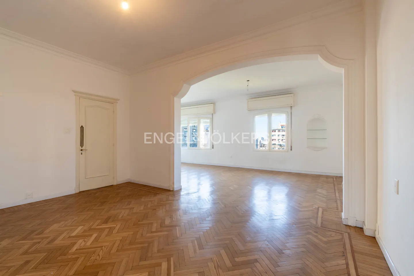 Bright, empty room with herringbone wood floors, white walls, arched doorway, and windows with city views.