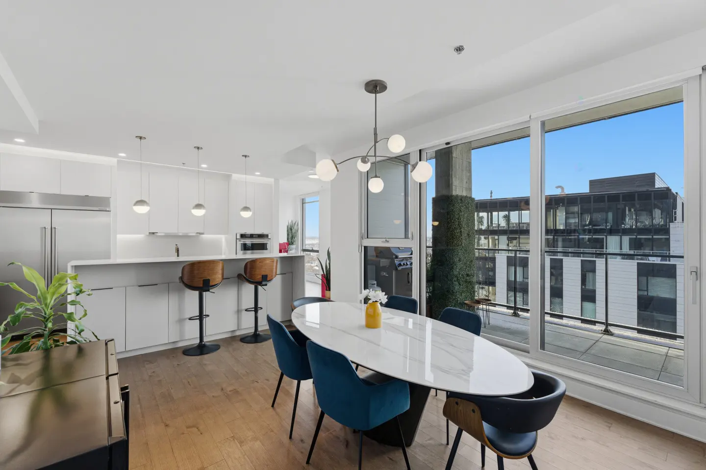 Bright, modern apartment interior with white kitchen, dining table with blue chairs, and large windows with city views.