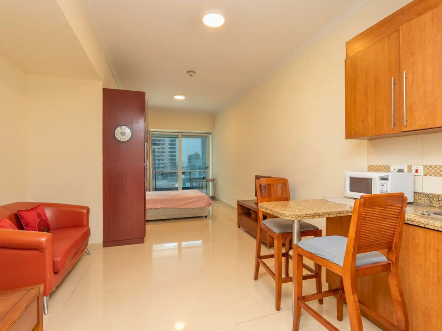 Studio apartment interior with a red sofa, wooden cabinets, a table with two chairs, and a bed visible through an open doorway.