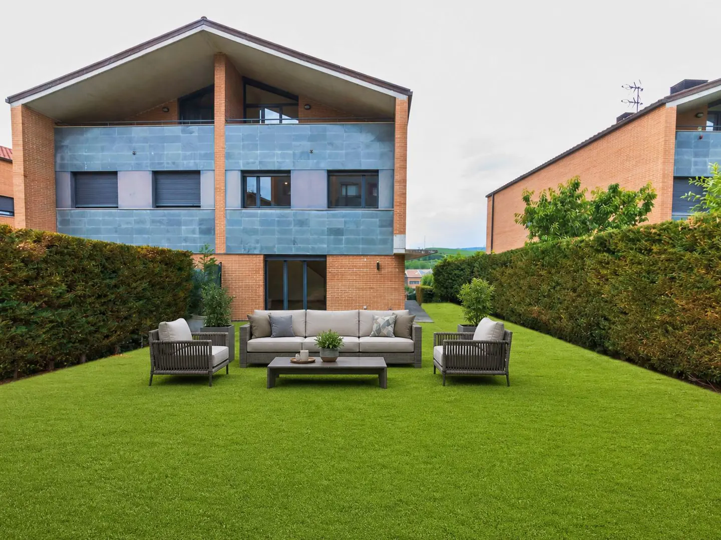 Backyard with green grass, a gray sofa, two chairs, and a coffee table in front of a brick and blue-tiled house.
