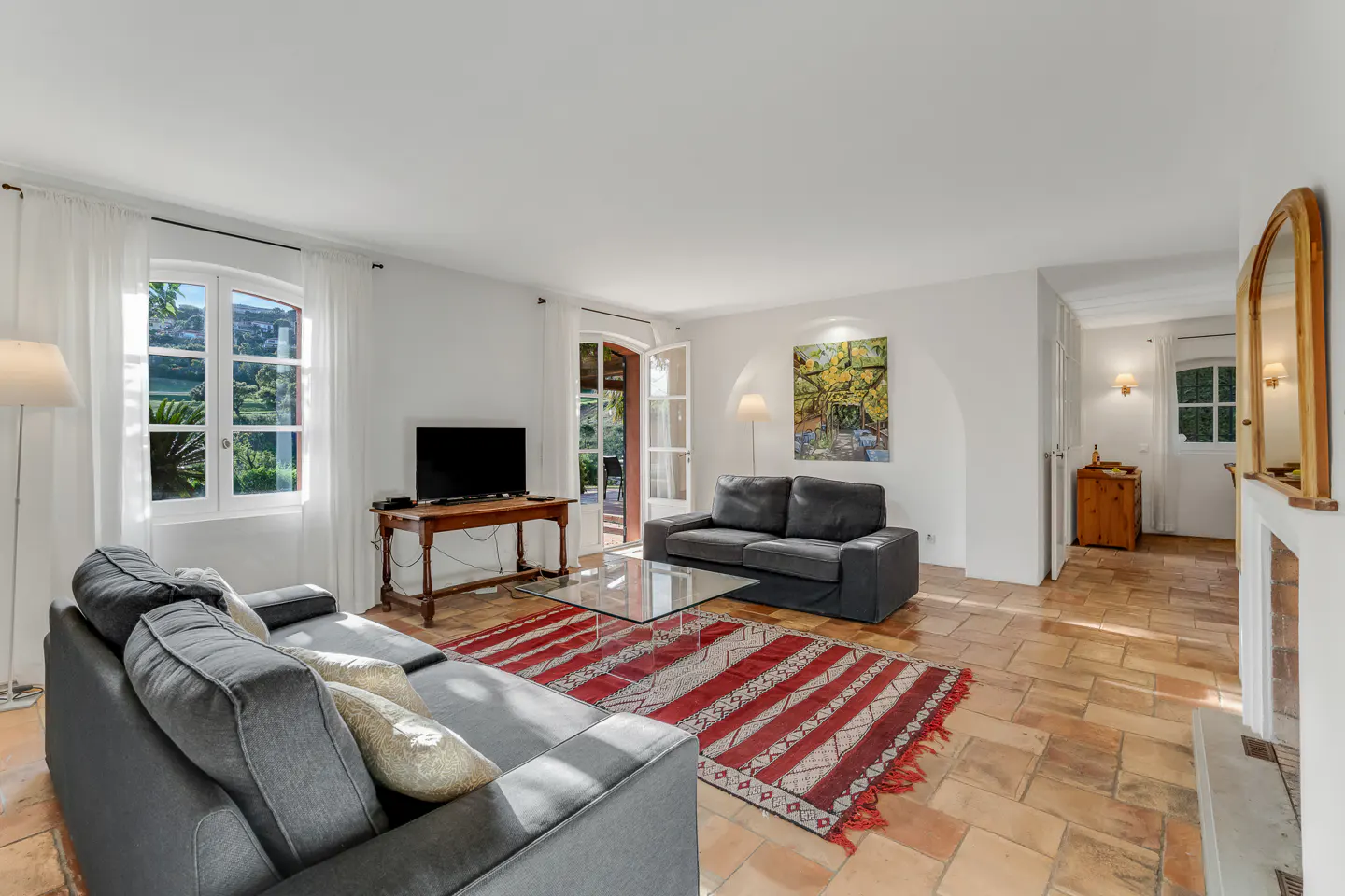 Bright living room with terracotta tile floor, gray sofas, and a red and white striped rug. A TV sits on a wooden table. French doors lead to a patio.