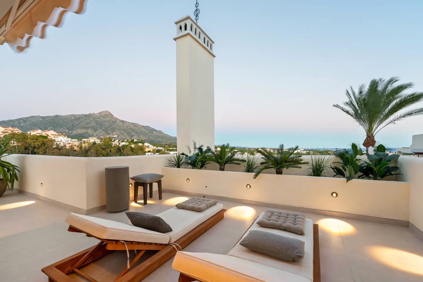 Rooftop patio with two lounge chairs, pillows, and a small table. A tall chimney and mountain views are in the background.