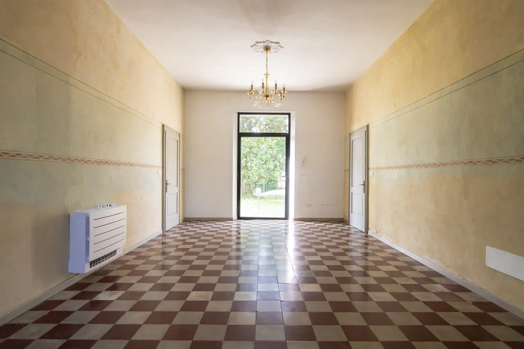 A long, empty room with a checkered floor, cream and yellow walls, and a chandelier.