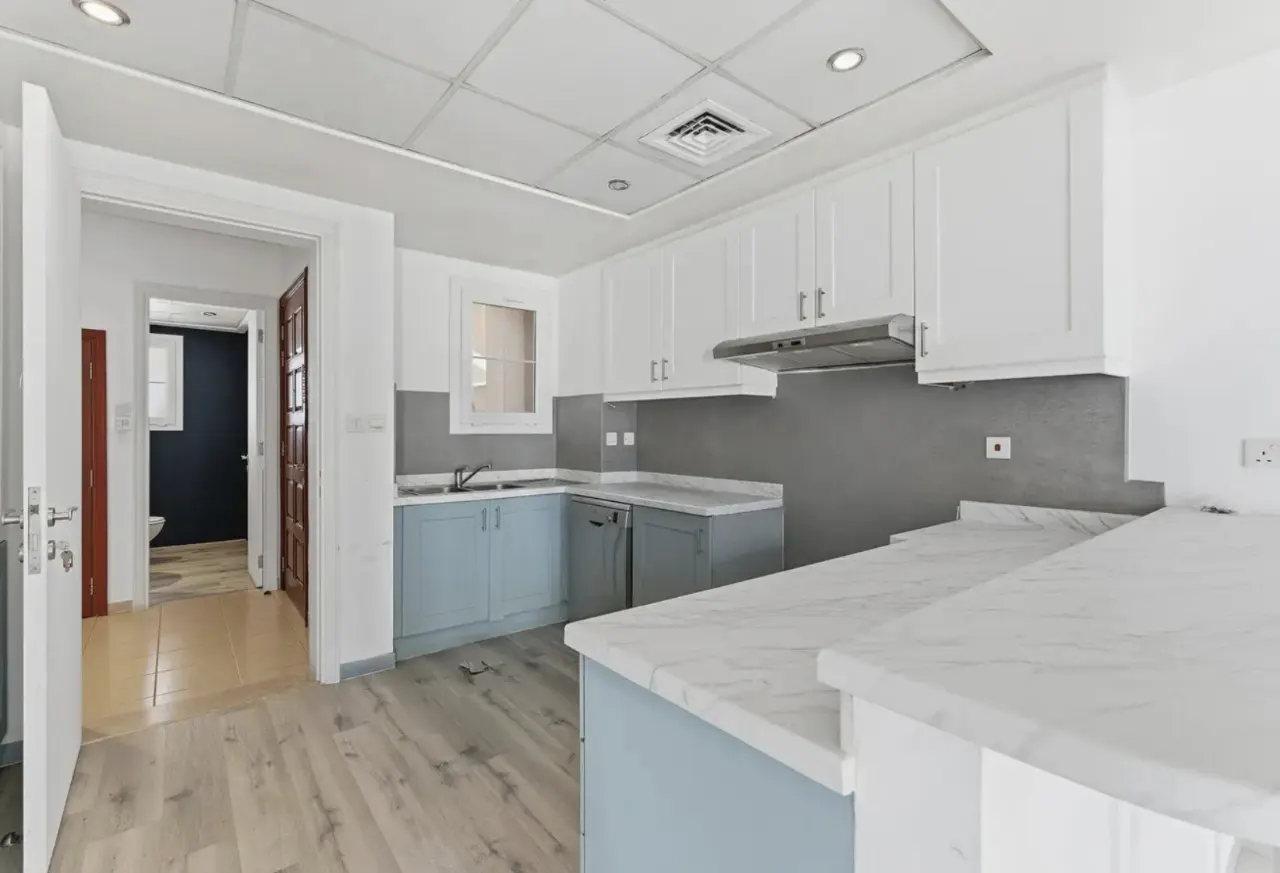 A bright kitchen with white cabinets, blue lower cabinets, and marble countertops. A doorway leads to a bathroom.