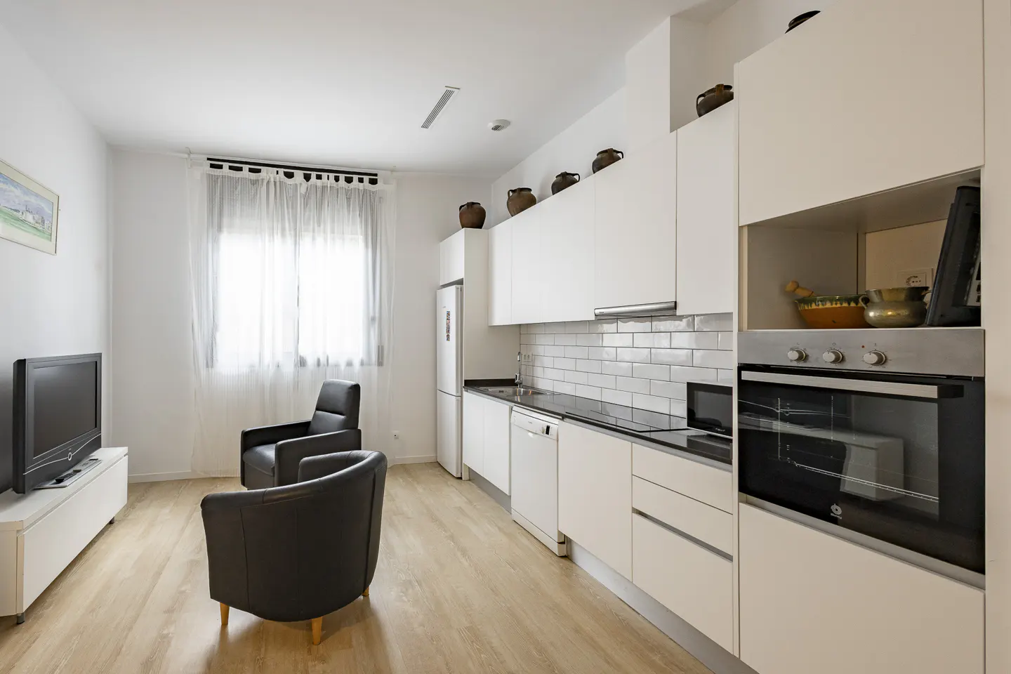 Modern kitchen with white cabinets, subway tile backsplash, and stainless steel oven, plus two black armchairs and a TV.