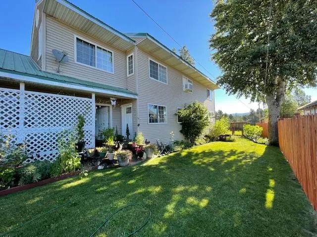Two-story beige house with a green metal roof, white lattice porch, potted plants, and a large grassy backyard.