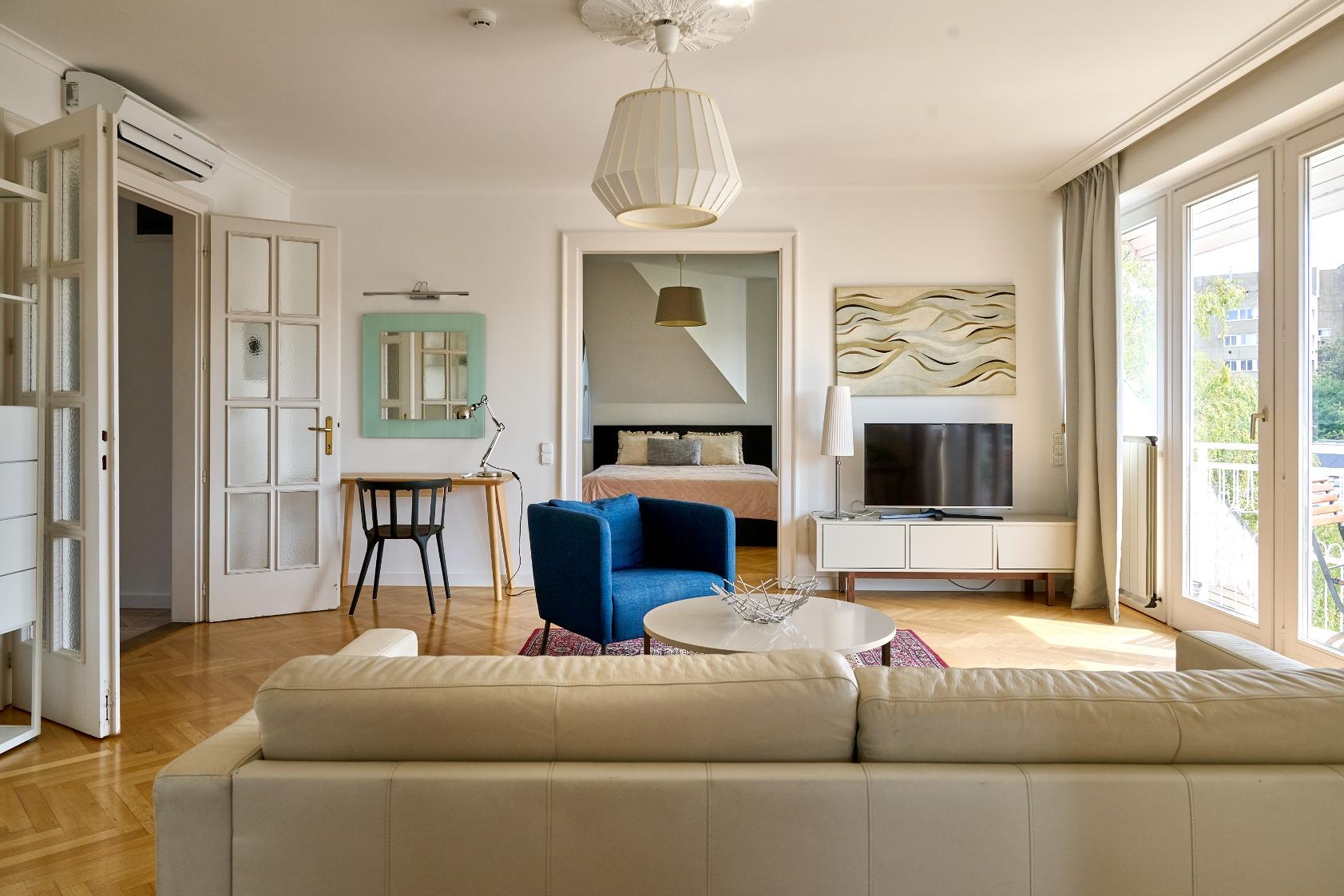 Bright living room with a beige sofa, blue armchair, round coffee table, and a view into a bedroom with a double bed.