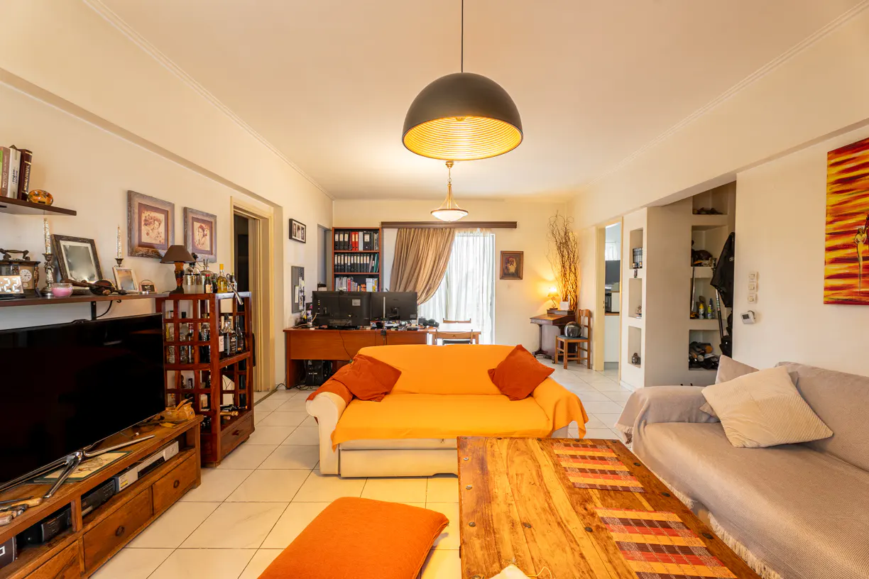 Living room with orange sofa, wood coffee table, TV, and shelves. A desk with monitors is visible in the background.