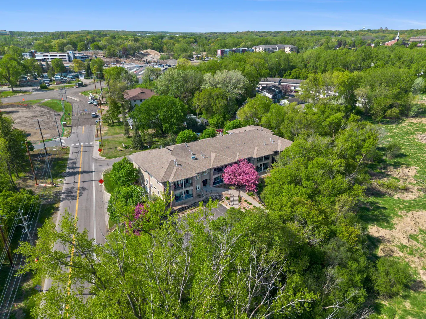 Aerial view of a two-story apartment building with a brown roof, surrounded by green trees and a pink flowering tree. A road runs alongside the building.