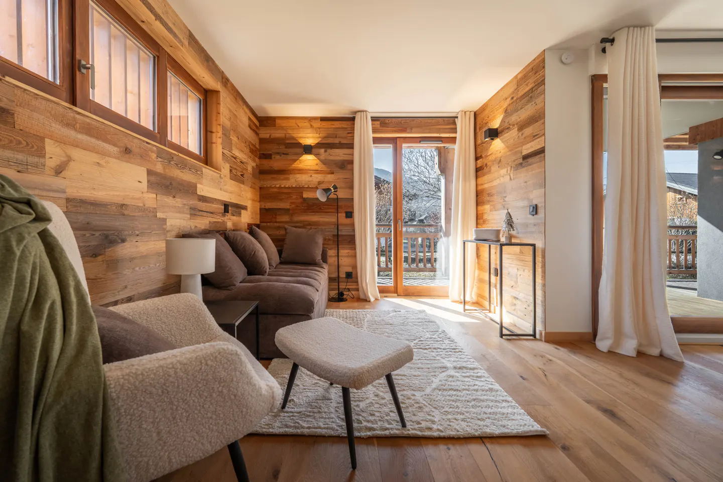 Cozy living room with wood paneled walls, hardwood floors, and a brown sofa. Balcony doors open to a snowy mountain view.