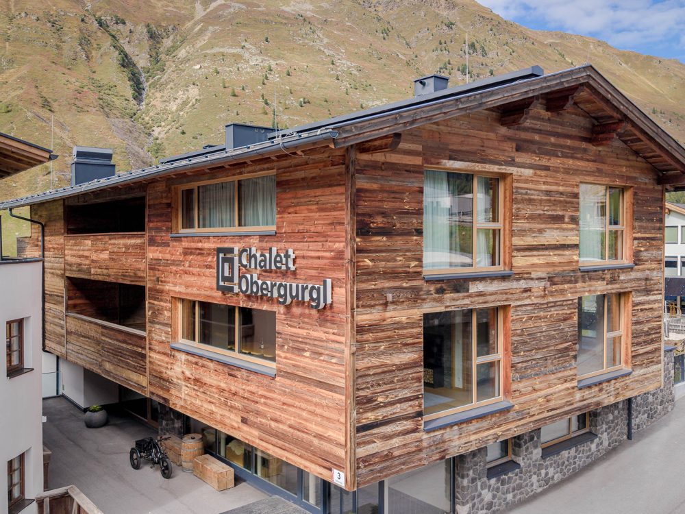 Exterior view of a rustic wooden Chalet Obergurgl building set against a backdrop of steep, grassy mountain slopes. [AI-generated text]