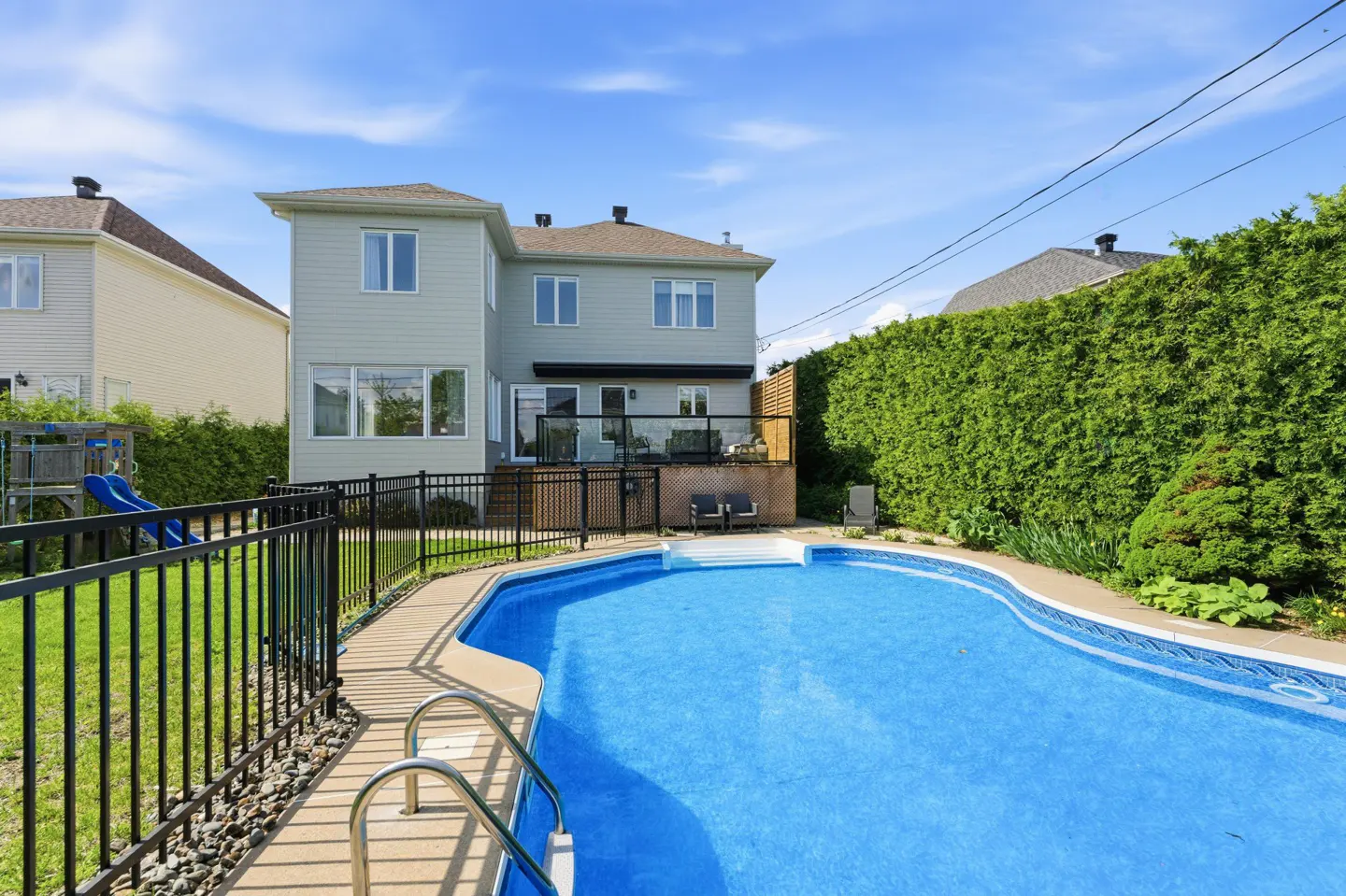 Backyard view of a two-story house with a blue swimming pool, deck, black fence, and green hedges.