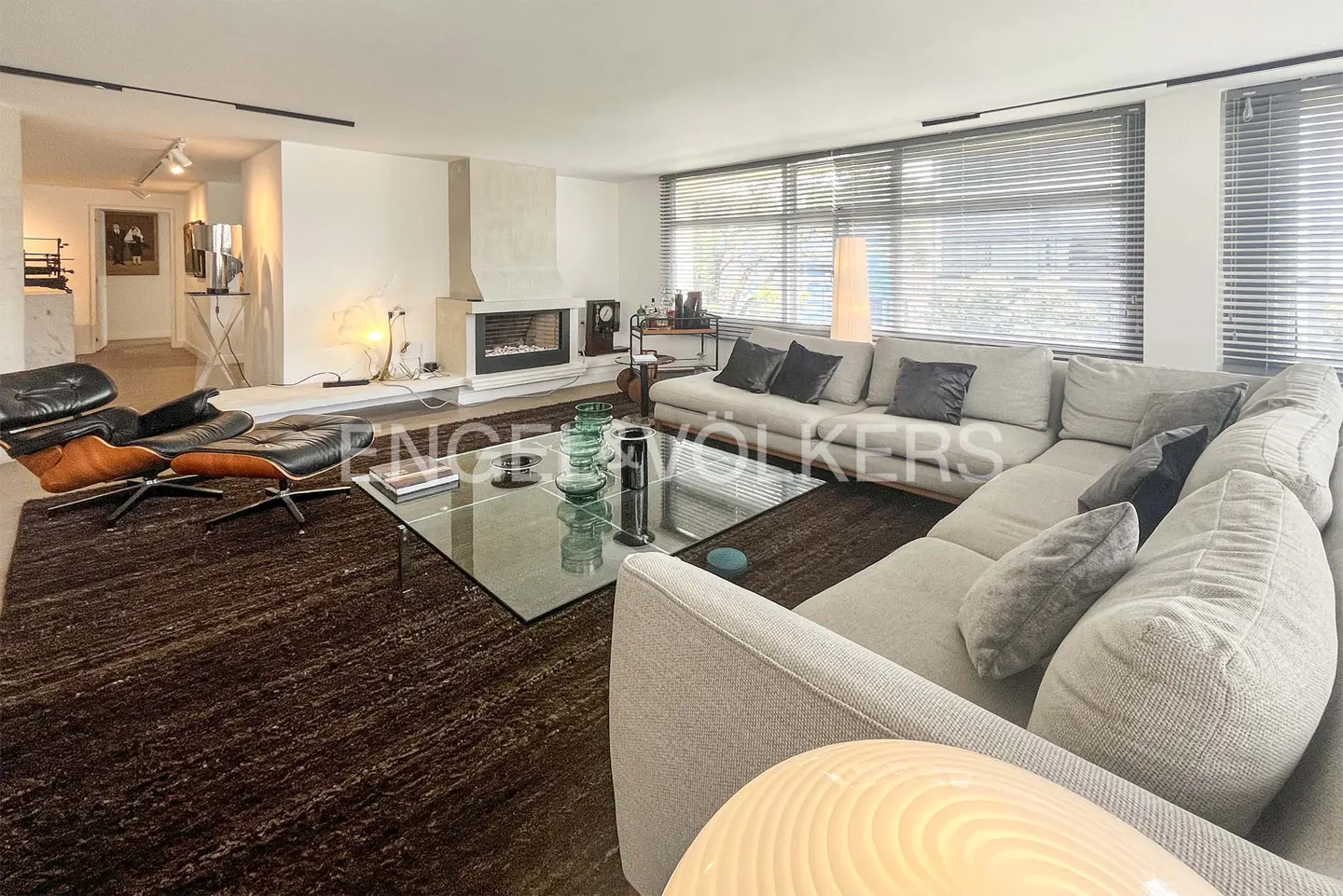 A modern living room with a large gray sectional sofa, glass coffee table, and brown rug. A fireplace and Eames chair are also visible.