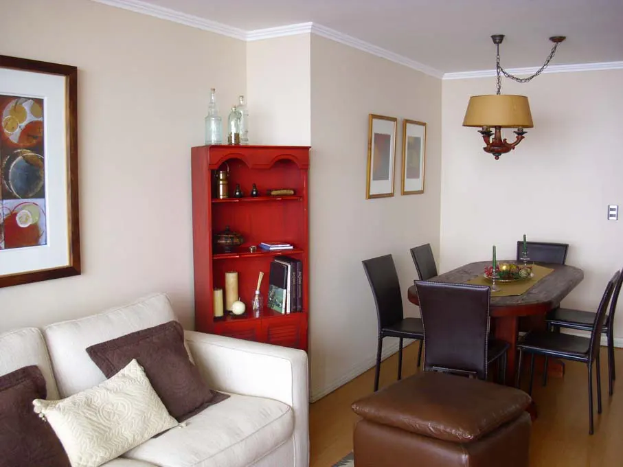 Living and dining area with a white sofa, brown pillows, red bookcase, and a dining table with black chairs. A tan lampshade hangs above.