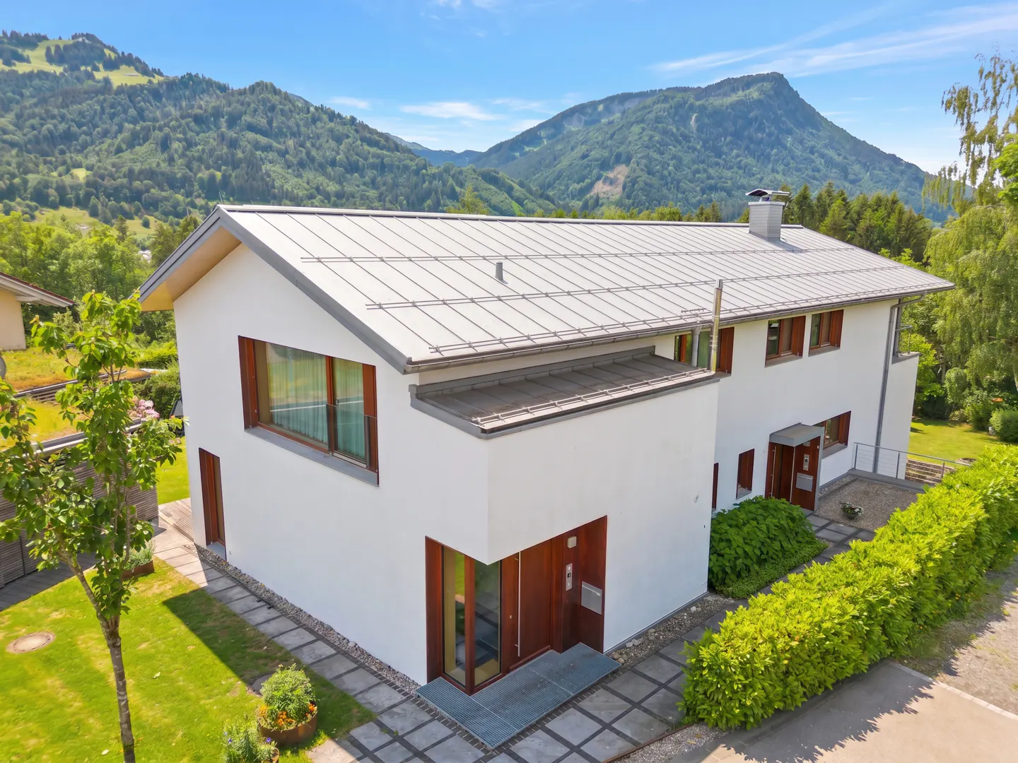 Modern white two-story house with a metal roof, surrounded by a green lawn and hedges, set against a mountain backdrop.